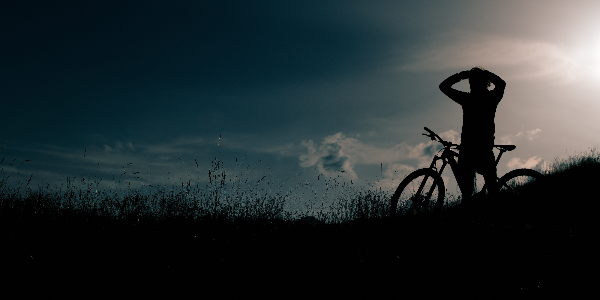 Silhouette eines Radfahrers, der am Hang steht, mit einem Fahrrad neben sich, bei Sonnenuntergang.