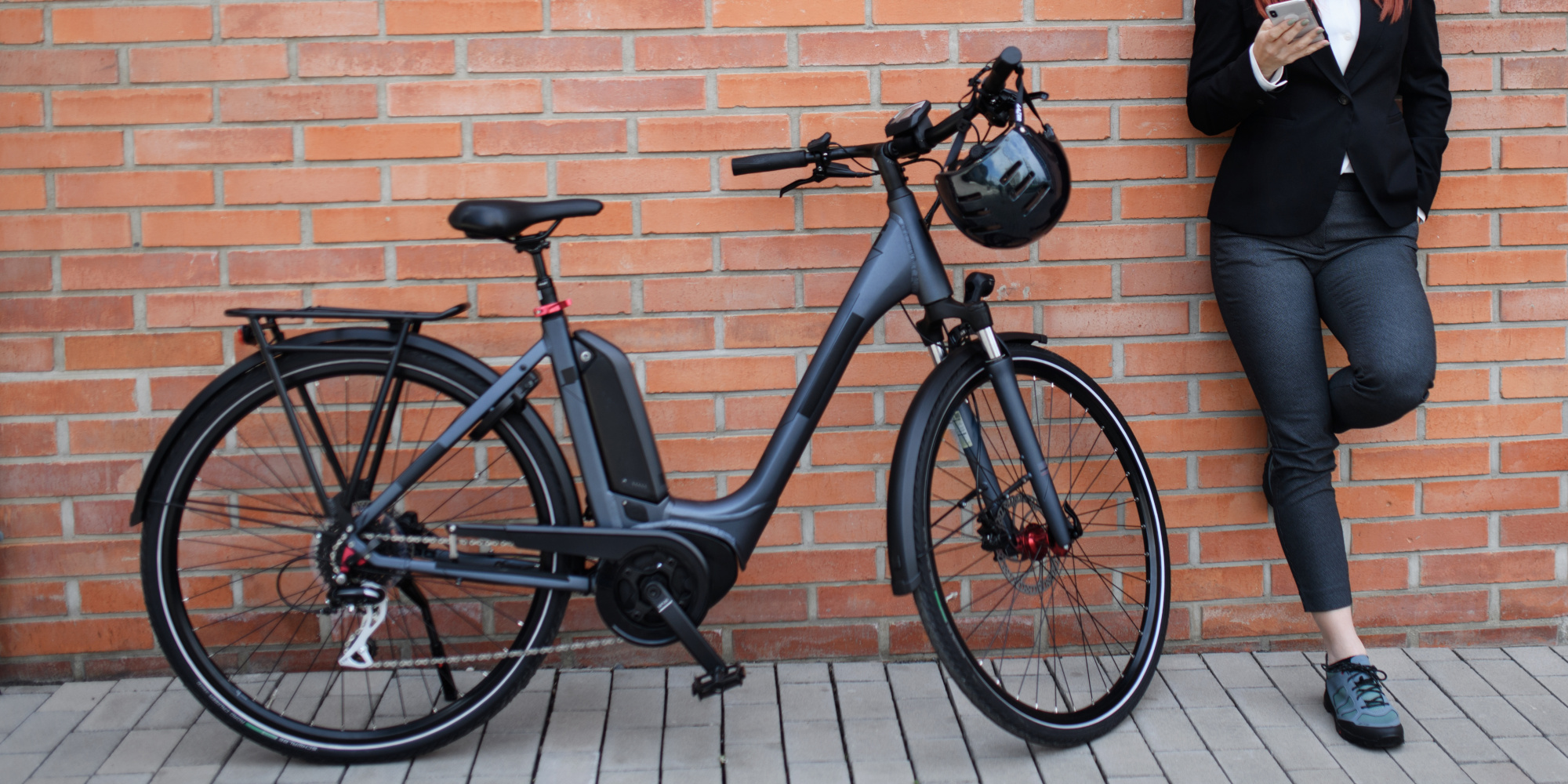 Eine Person steht neben einem schwarzen Elektrofahrrad vor einer Ziegelsteinwand.