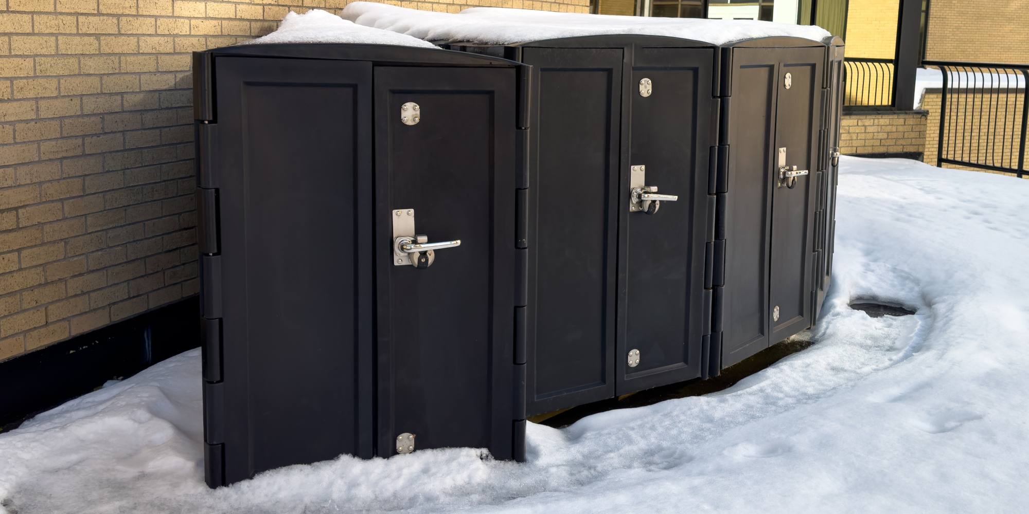 Vier schwarze Abfallbehälter stehen in einer Schneeoberfläche neben einem Gebäude.