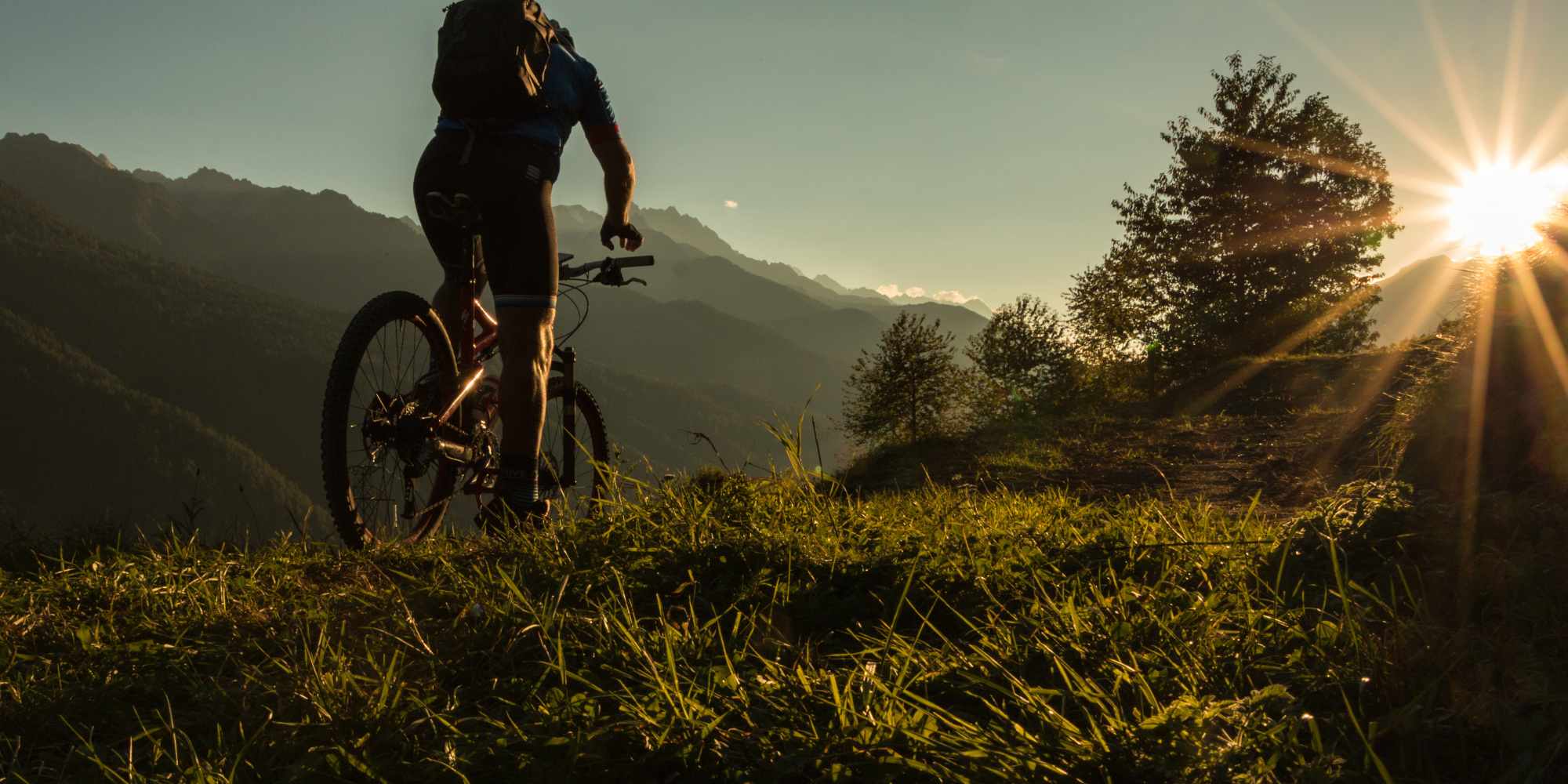 Ein Radfahrer steht auf einer Wiese mit Bergen und Sonnenlicht im Hintergrund.