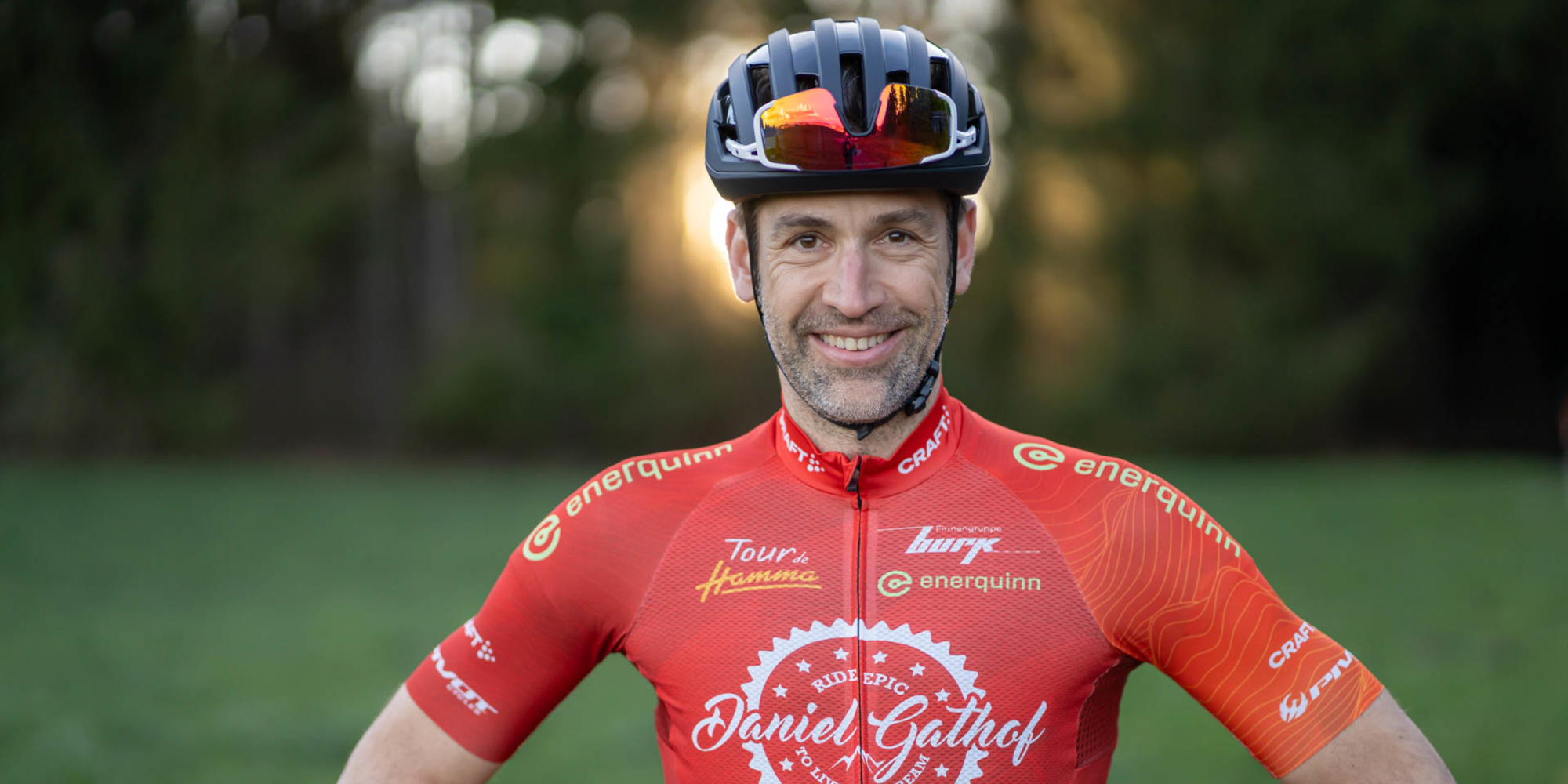 Ein Radfahrer in einem leuchtend roten Trikot steht im Freien, umgeben von Bäumen.