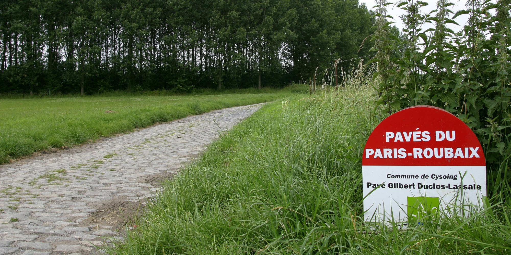 Pflasterweg mit grünem Gras und einem Schild zum Paris-Roubaix-Radrennen.