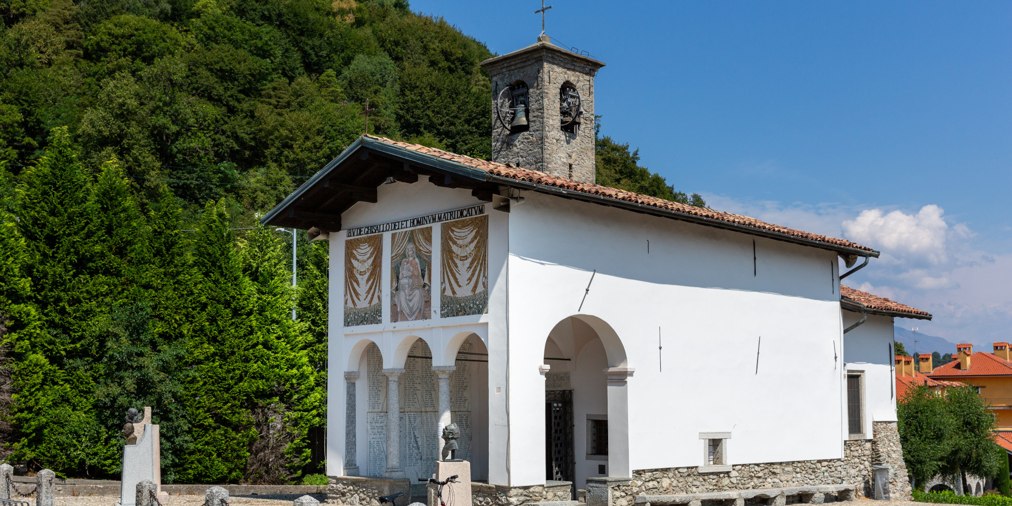 Weißes, traditionelles Gebäude mit Fresken und Glockenturm, umgeben von grünen Bäumen.