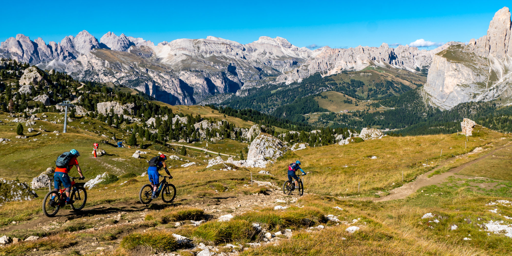 Mountainbiker fahren auf einem Pfad durch eine alpine Landschaft mit Bergen im Hintergrund.