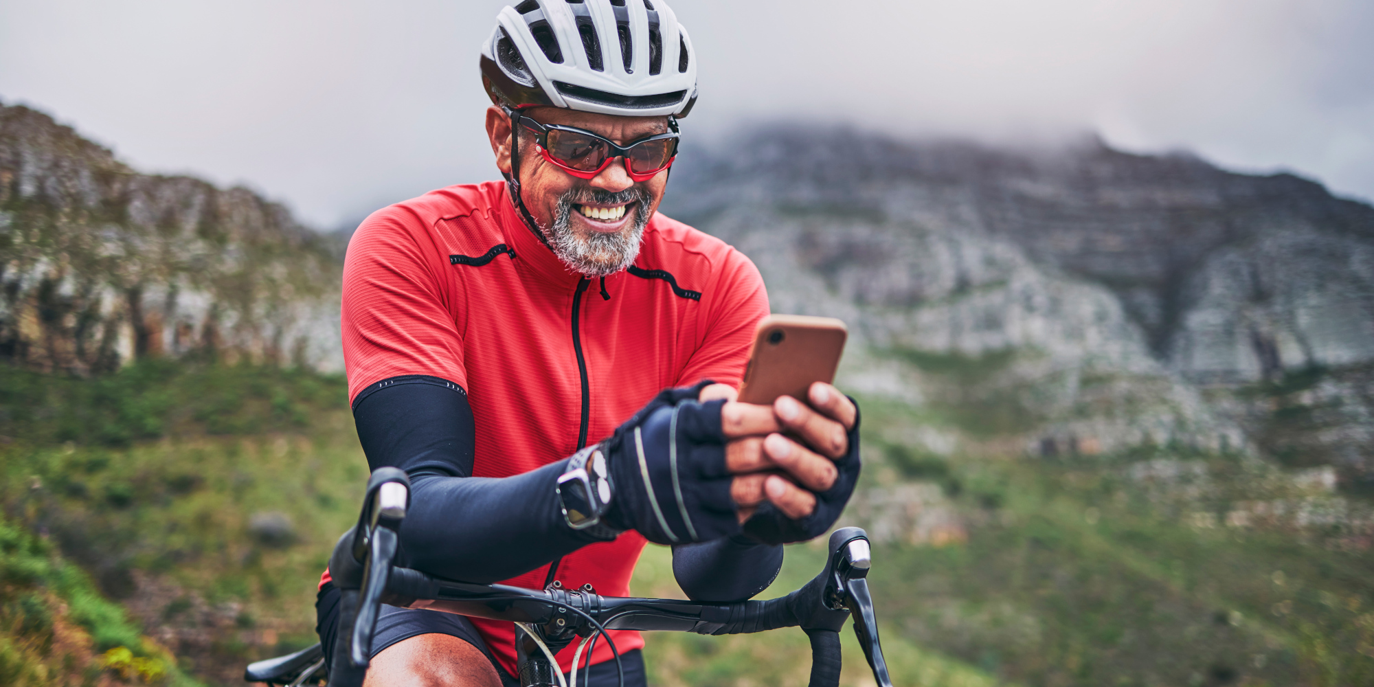Radfahrer in rotem Trikot, der am Handy sitzt, mit bergiger Landschaft im Hintergrund.