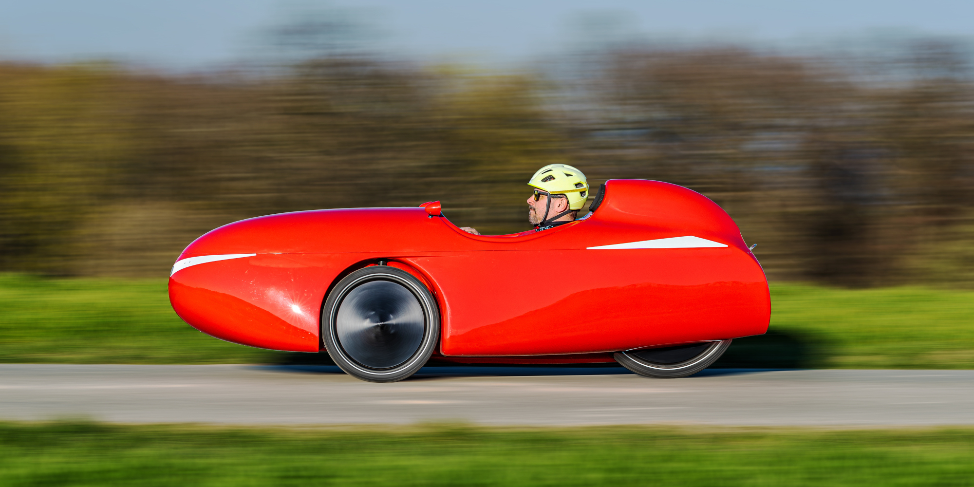 Entdecke alle Vor- und Nachteile des Velomobils Ein Mann fährt in einem roten Velomobil auf einer Straße