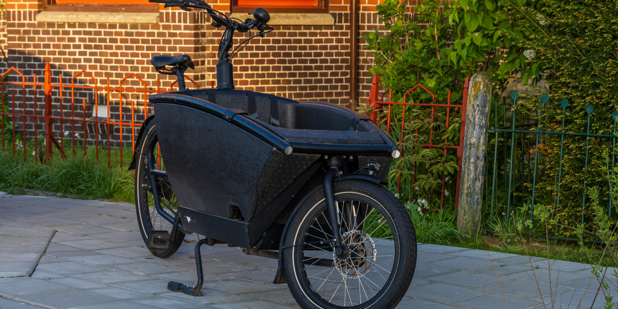 Schwarzes Lastenrad steht auf einem gepflasterten Weg vor einem Garten.