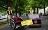 Zwei Personen in leuchtenden Westen stehen neben einem Fahrrad-Rickshaw in einem Park.