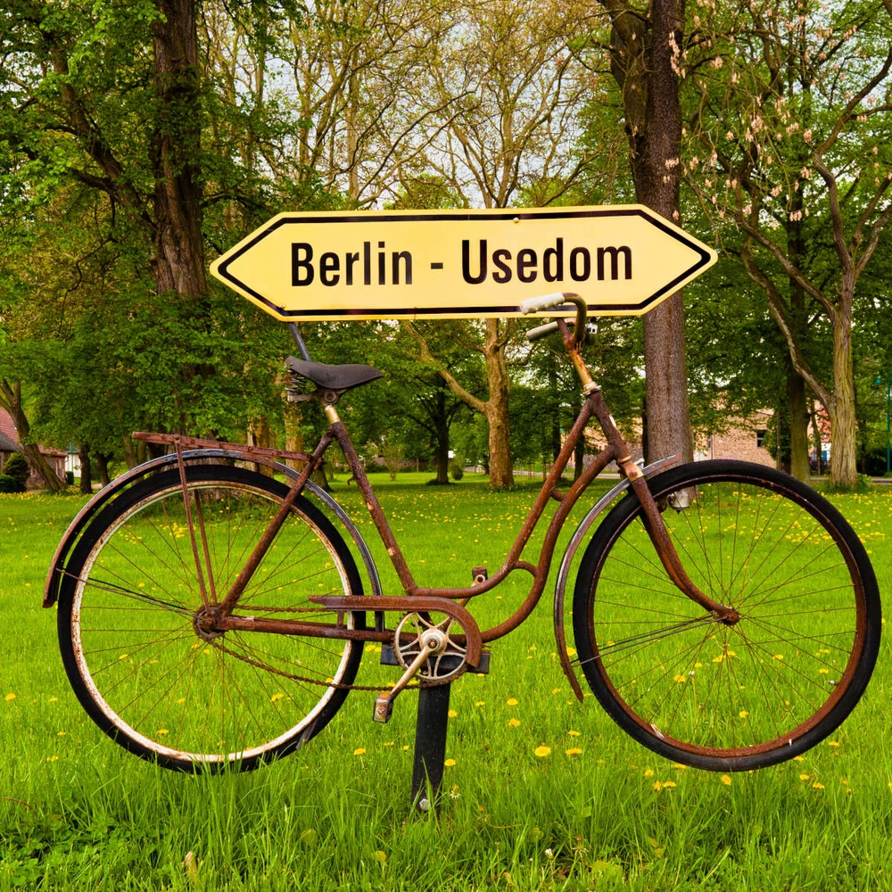 Ein rostiges Fahrrad steht neben einem Wegweiser nach Berlin und Ueckermünde in einer grünen Wiese.