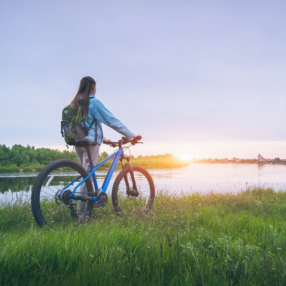 Frau fährt auf Dirt Bike durchs Grün mit Blick auf den Sonnenuntergang am Seeufer