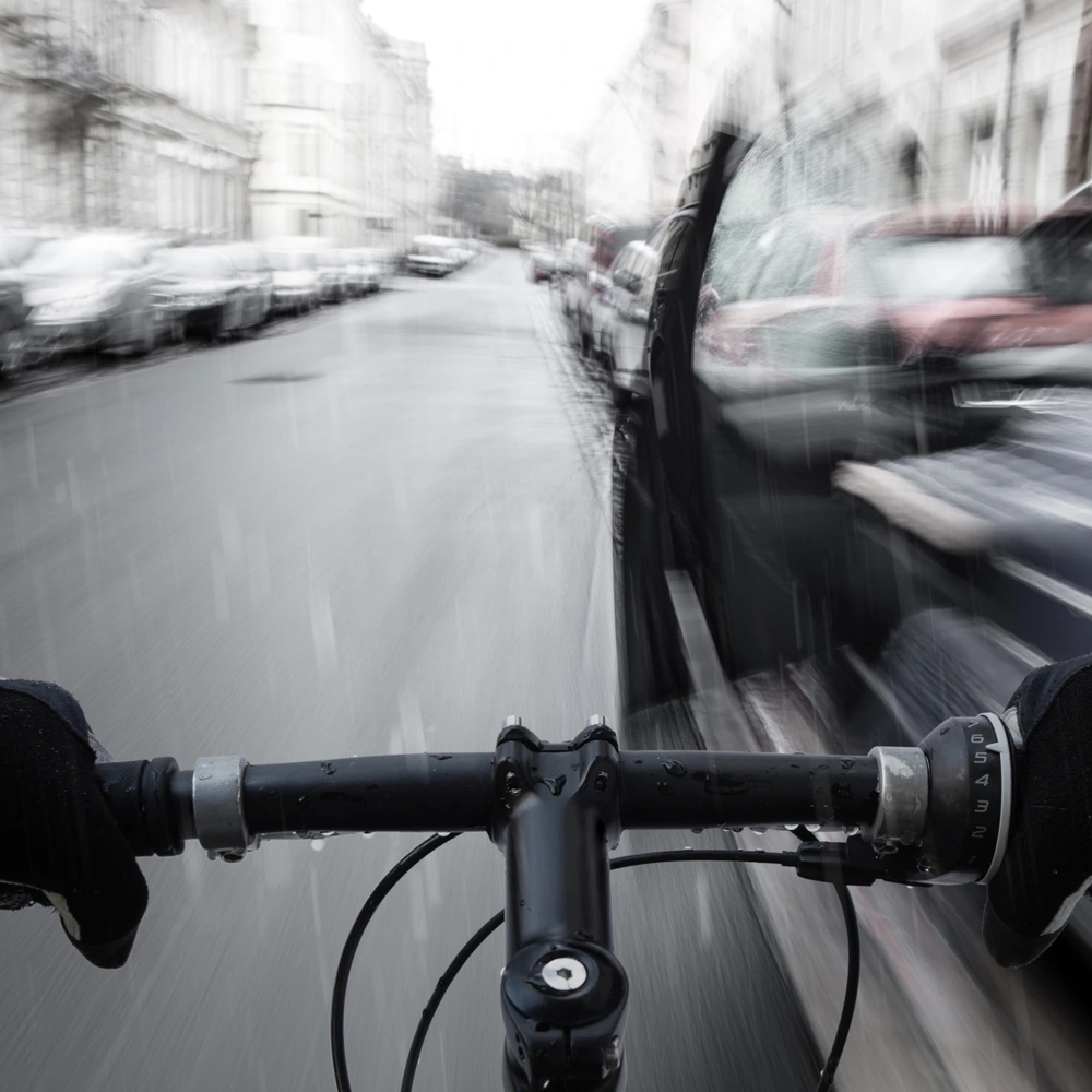 Blick von einem Fahrradlenker auf eine verregnete Straße mit vorbeirasenden Autos.