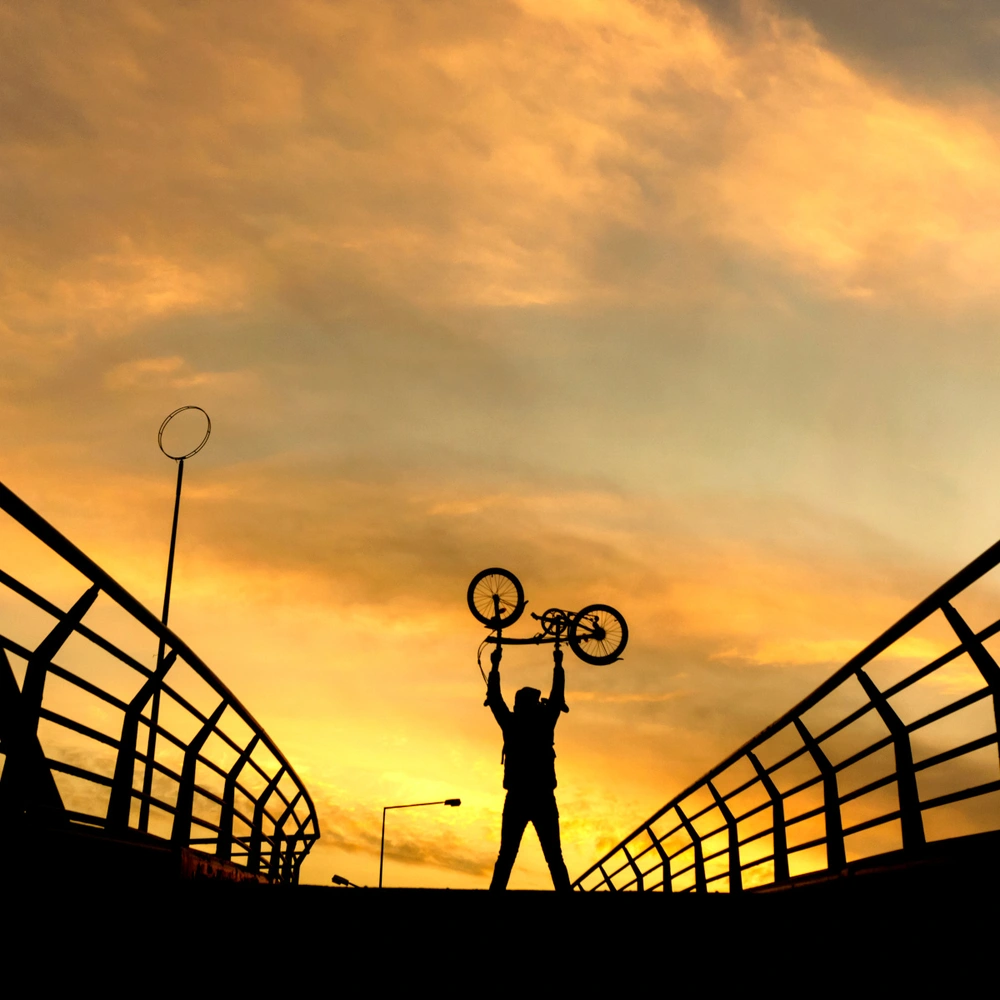 Mann auf einer Brücke hält ein Faltrad in die Luft im Sonnenuntergang
