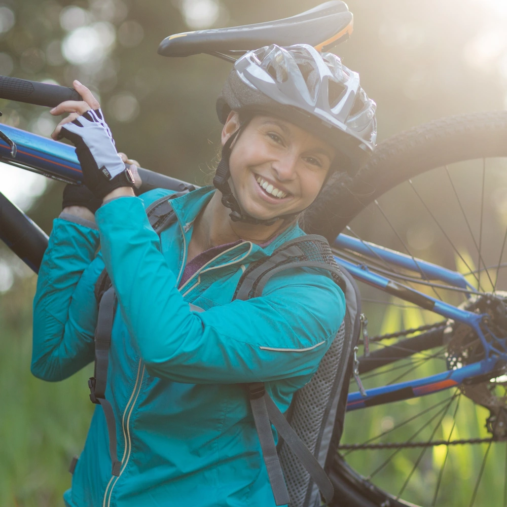 Lächelnde Person trägt ein Mountainbike in einer sonnigen, grünen Landschaft.