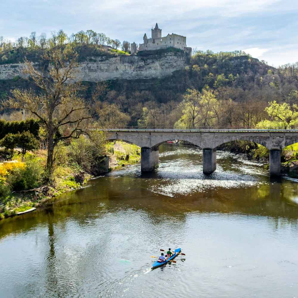 Flusslandschaft mit Ruderboot, Brücke und Anwesen unter einer Burgruine.