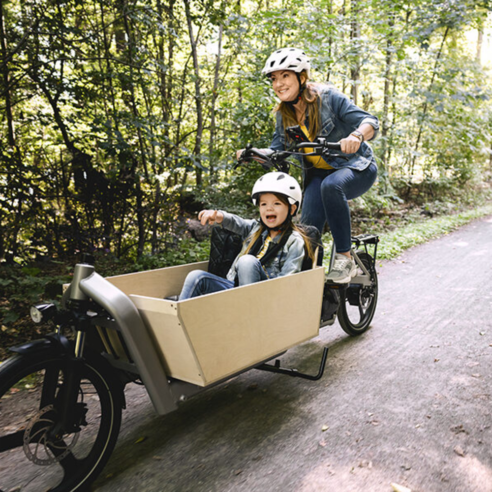 Diese Risiken sollten Sie bei der Fahrt mit einem Lastenrad beachten. Frau fährt mit Kind und Helm auf Lastenrad durch den Wald