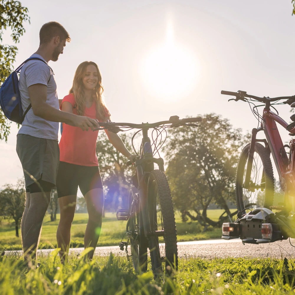 Paar mit Fahrrädern vor einem Auto, umgeben von Bäumen im Sonnenschein.