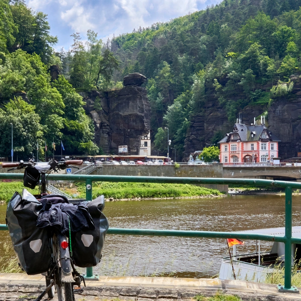 Fahrrad mit Gepäck nahe einem Fluss, umgeben von grünen Bergen und einem kleinen Ort.