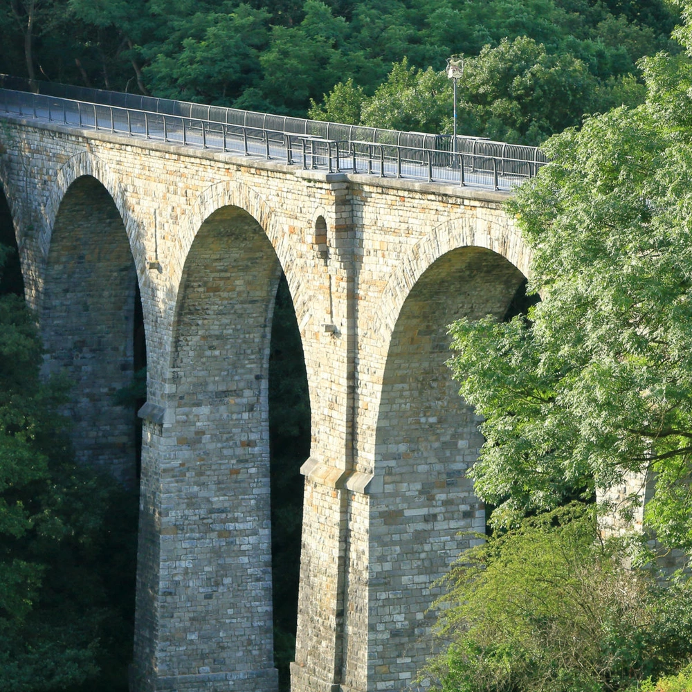 Steinernes Viadukt mit mehreren Bögen, umgeben von Bäumen und Grün.