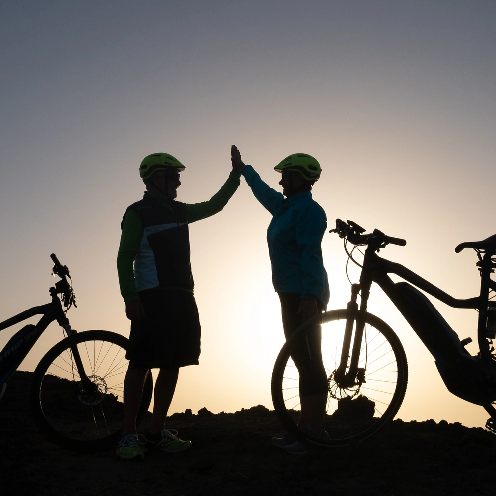 Zwei Personen stehen neben ihren E-Bikes vor dem Sonnenuntergang und klatschen sich ab