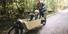 Diese Risiken sollten Sie bei der Fahrt mit einem Lastenrad beachten. Frau fährt mit Kind und Helm auf Lastenrad durch den Wald