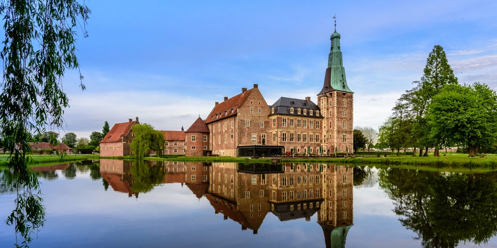 Historisches Schloss mit Turm, umgeben von Bäumen, gespiegelt im ruhigen Wasser.