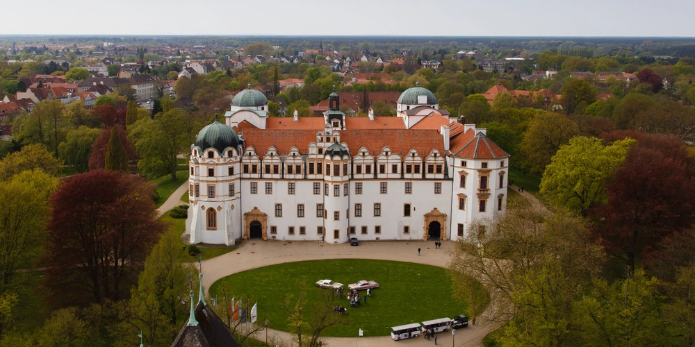 Luftaufnahme eines historischen Schlosses mit roten Dächern und umgebendem grünen Park.