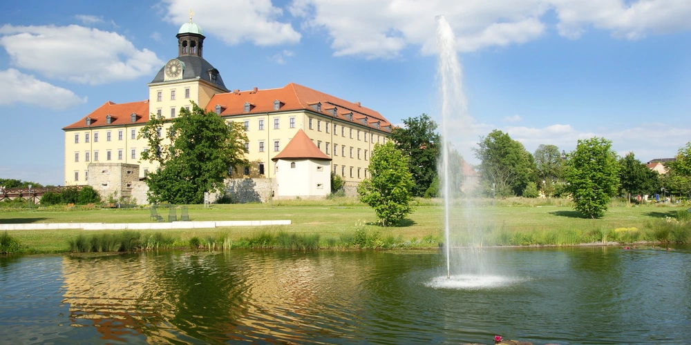 Schloss mit roten Dächern und Brunnen im Vordergrund, umgeben von Grünflächen.