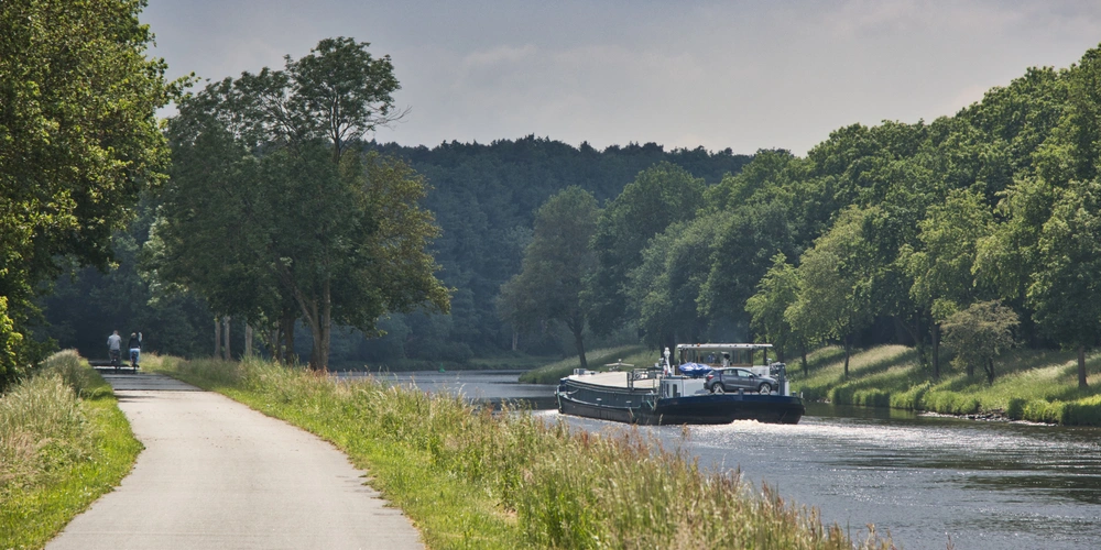 Ein Frachtschiff fährt auf einem ruhigen Kanal neben einem Radweg und Bäumen.