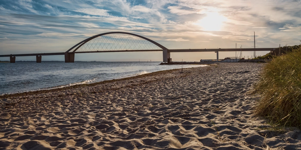 Brücke über das Wasser bei Sonnenuntergang, Sandstrand im Vordergrund.