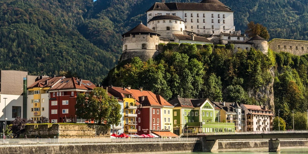 Blick auf eine Festung mit bunten Häusern am Flussufer, umgeben von Bergen