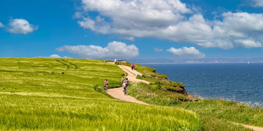 Grüner Hügelweg mit Spaziergängern und Blick auf das Meer unter blauem Himmel.
