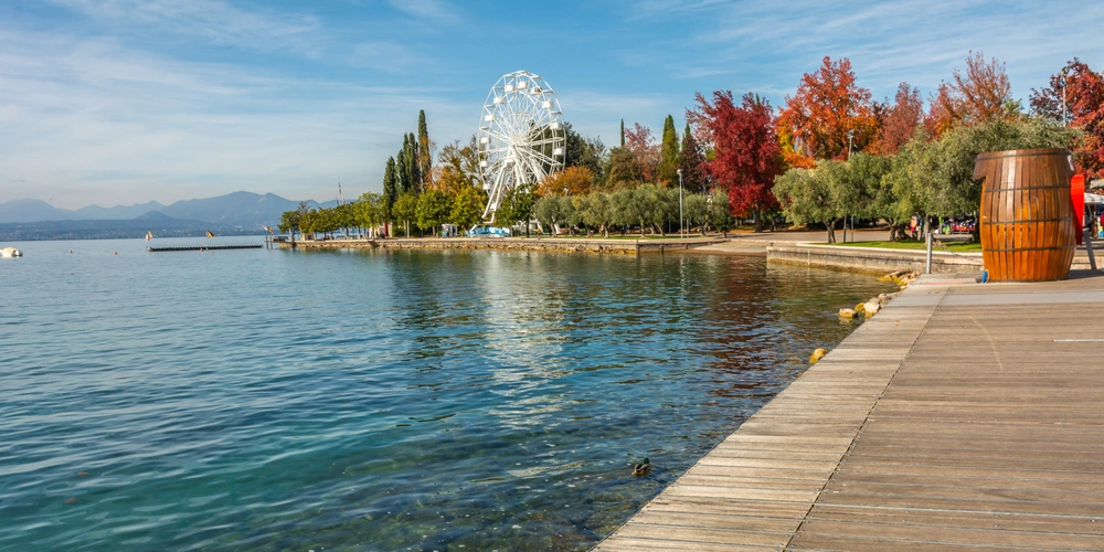 Ruhevoller See mit farbenfrohen Bäumen und Riesenrad im Hintergrund.