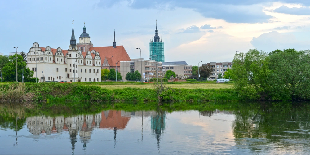 Historische Gebäude mit Türmen spiegeln sich im ruhigen Wasser eines Flusses.