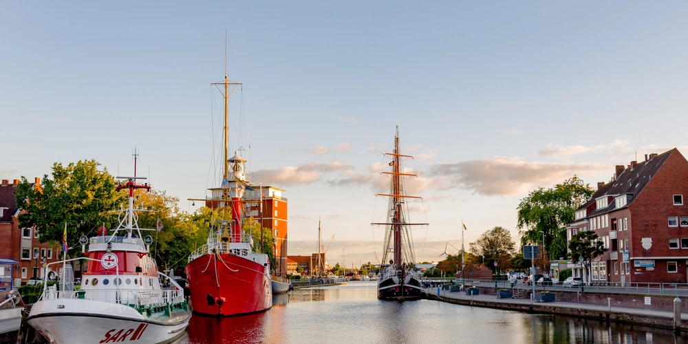 Hafenansicht mit roten Schiffen und einem historischen Segelschiff im Abendlicht.