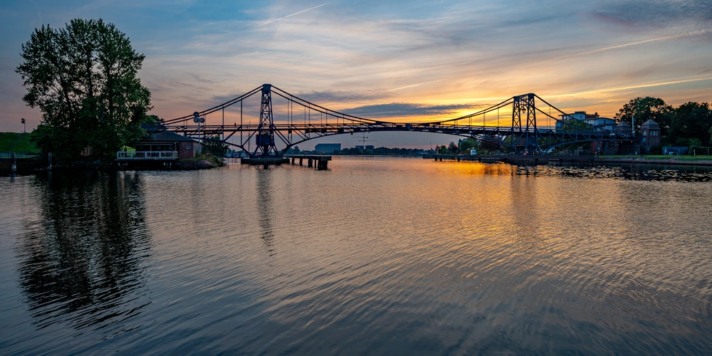 Blick auf eine Brücke bei Sonnenuntergang, gespiegelt im ruhigen Wasser.