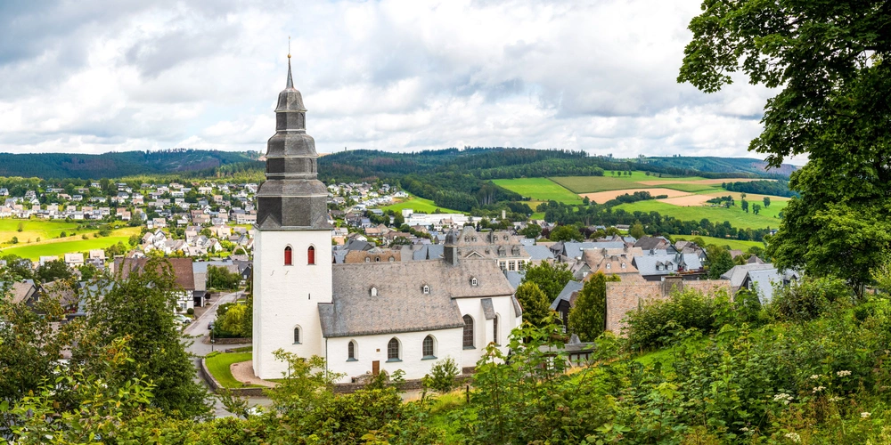 Weißes Kirchturmgebäude mit Spitze umgeben von grünen Feldern und Hügeln.