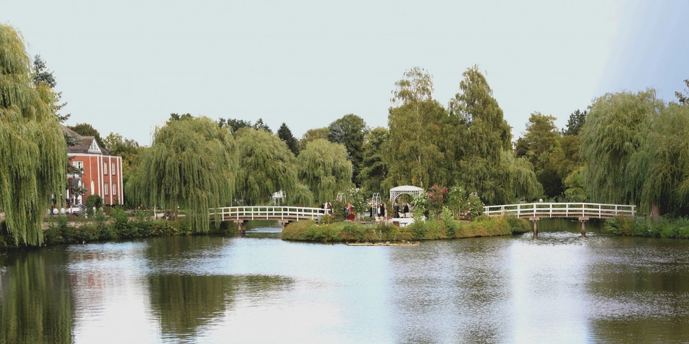 Idyllische Seenlandschaft mit Bäumen, einer Brücke und einem Pavillon im Hintergrund.