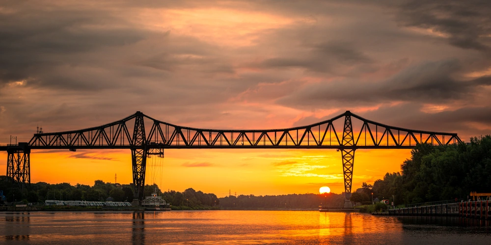 Stahlbrücke im Sonnenuntergang, reflektiert im ruhigen Wasser.