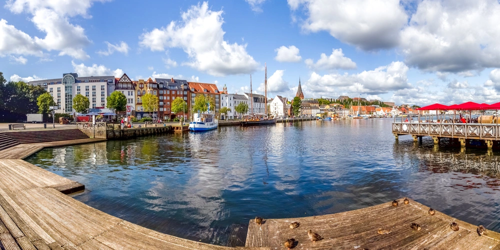 Hafenansicht mit Booten, Holzsteg und bunten Gebäuden unter blauem Himmel.