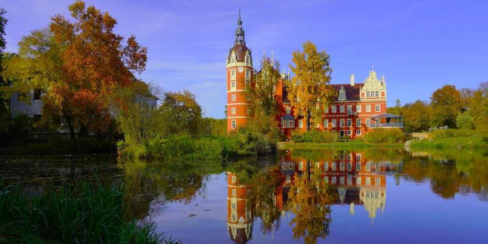 Schloss am Ufer eines ruhigen Sees, umgeben von buntem Herbstlaub und klarem Himmel.
