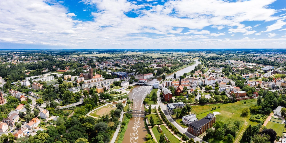 Luftaufnahme einer Stadt mit Fluss, grünen Flächen und modernen Gebäuden unter blauem Himmel.