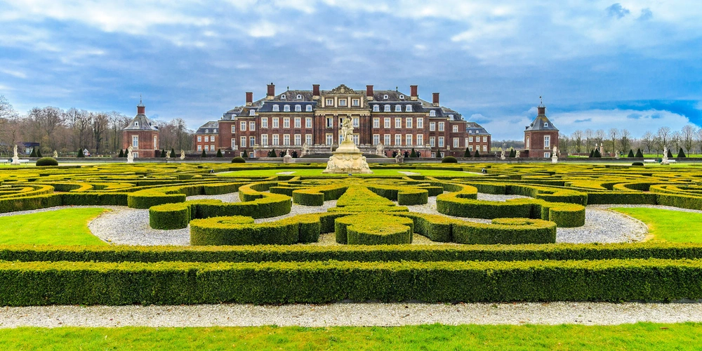 Schloss mit gepflegtem Garten und geometrischen Hecken, unter einem bewölkten Himmel.