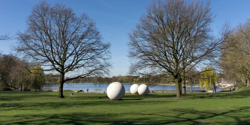 Grüner Park mit drei weißen, eiförmigen Skulpturen und Bäumen am Ufer eines Gewässer.