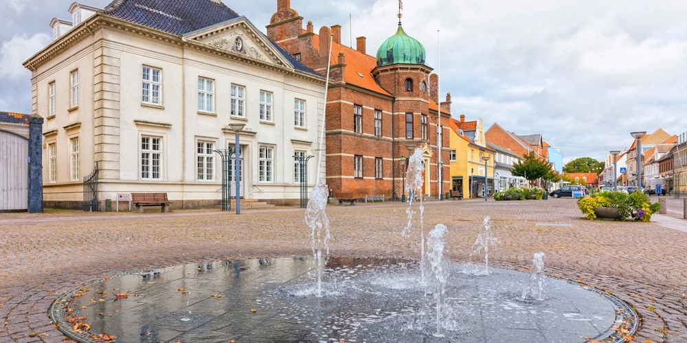 Brunnen in einer Stadt mit historischen Gebäuden und bewölktem Himmel.
