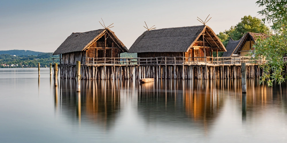 Drei traditionelle Holzhäuser auf Stelzen am ruhigen Wasser, sanfte Farben im Hintergrund.