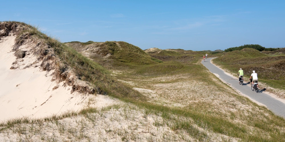 Radfahrer auf einem Weg durch sandige Dünenlandschaft unter blauem Himmel.