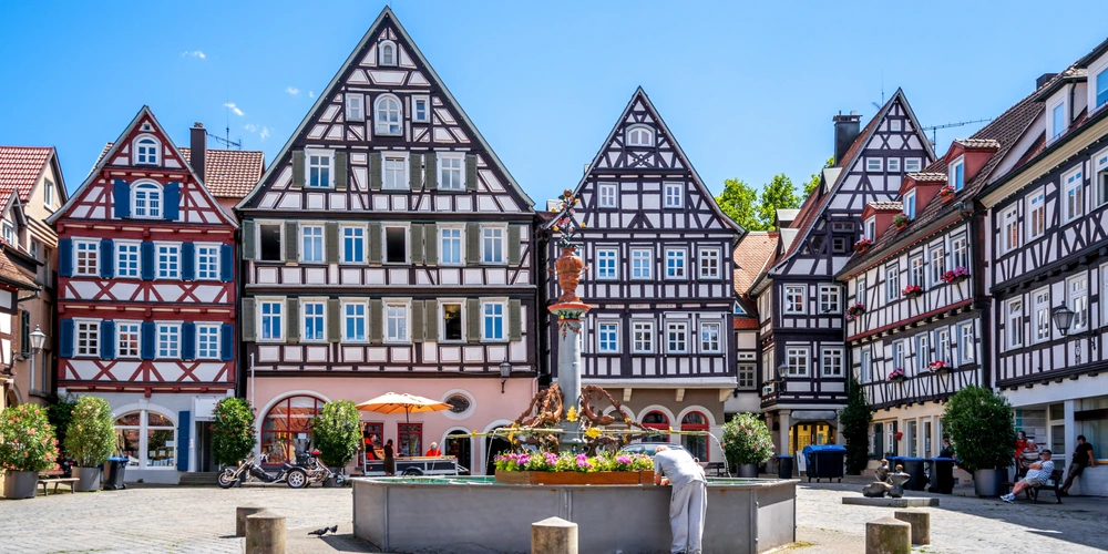 Fachwerkhäuser um einen zentralen Brunnen auf einem sonnigen Platz.