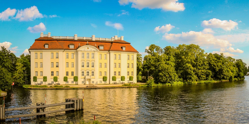 Historisches Gebäude am Wasser, umgeben von Bäumen und blauem Himmel mit Wolken.
