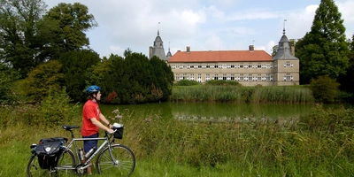 Radfahrer in rot vor einem historischen Gebäude an einem ruhigen Gewässer.