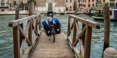 Radweg München-Venedig – Tipps für deine Alpentour Fahrrad mit Gepäck auf einem Holzsteg über dem Wasser, umgeben von Gebäuden.